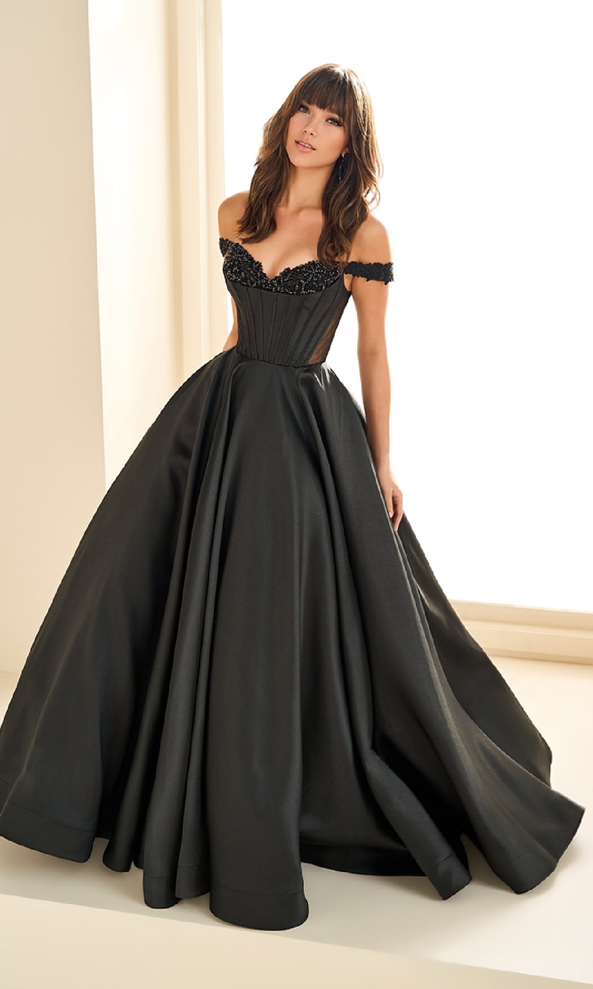A woman stands indoors wearing the Ellie Wilde EW36100, a long black prom dress featuring an off-the-shoulder neckline, fitted bodice, and dramatic full skirt.
